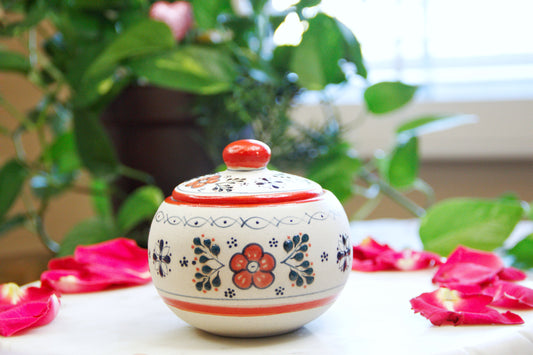 Front view of the handmade artisanal candle in a red floral design talavera with a closed lid on its side. Custom made by Artisan in Mexico. 100% All Natural Soy Candle. Reuse the talavera as home decor or storage.