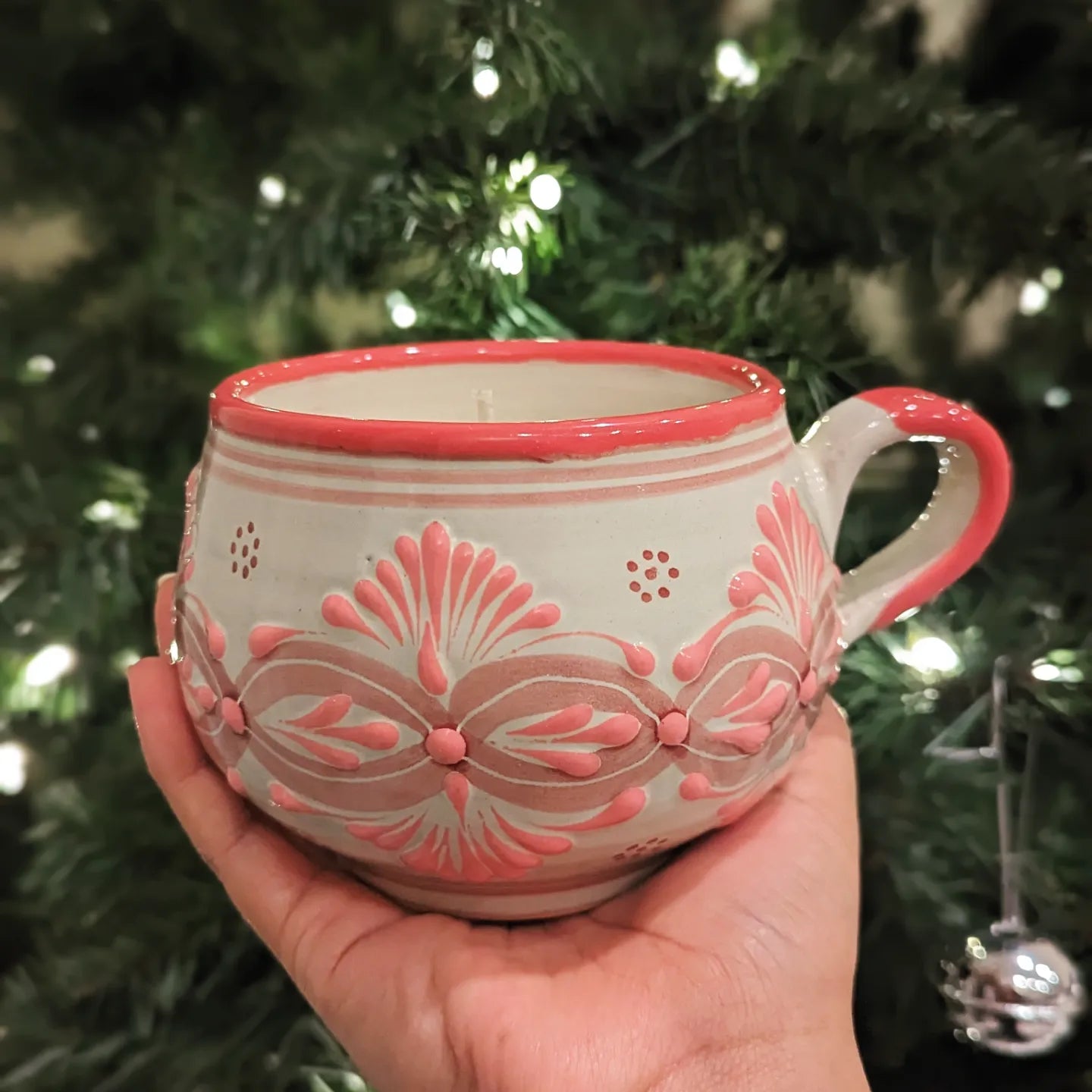 A hand holding an Artisanal candle in a beautiful light pink and biege handmade design talavera cup with a handle. Handcrafted by Artisan in Puebla, Mexico. 100% All Natural Soy Candle. Reuse the talavera as home decor or storage.